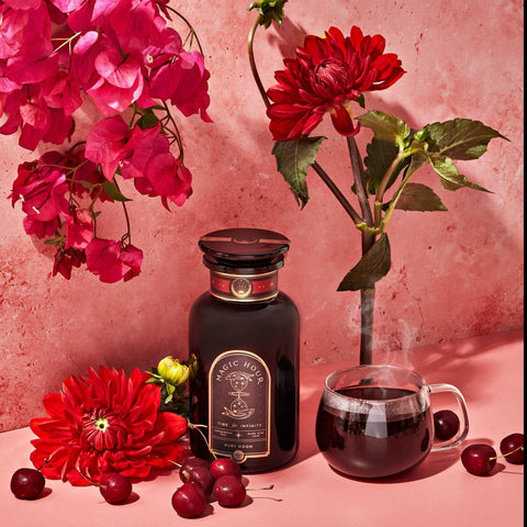 A vibrant still life scene features a bottle of Ruby Moon™ : Hibiscus Elderberry Tea by Club Magic Hour, a steaming transparent teacup, a bunch of fresh cherries, and bright red flowers against a pink backdrop. The arrangement highlights the rich red hues and the elegance of the tea setting.