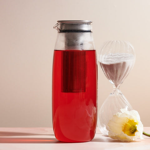 A Unitea Cold Brew Carafe filled with vibrant red liquid stands next to a delicate white flower and a clear hourglass with white sand. Featuring a built-in infuser basket, this carafe is an elegant piece of teaware. The neutral and minimalistic background highlights its modern design.