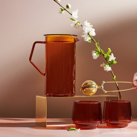 A still life composition featuring a tall Cast Sepia Pitcher, crafted from borosilicate glass, accompanied by two matching glasses and a brass tea infuser held by a hand. A delicate white blossom on a branch enhances the elegant and minimal aesthetic, set against a soft background.