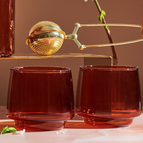 Two Sepia Tumblers from the Set of Four sit on a pink surface, their amber-colored glass evoking a vintage-inspired elegance. Above them, a gold tea infuser rests on a brass stand, with white petals scattered around. A delicate branch with small blossoms in the background adds to the warm and sophisticated scene.