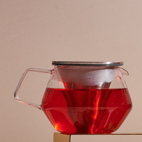 A Kinto Carat Teapot filled with rich red tea is shown against a beige background. The teapot, made of transparent glass, features a stainless steel lid and a handle on the left side. The vibrant color of the tea is beautifully visible through the clear glass.