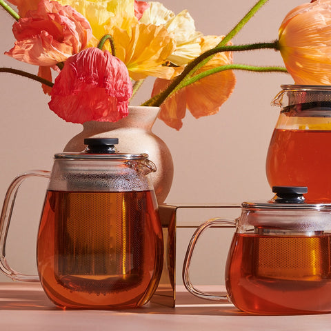 Two Magic Hour Kinto Stainless Steel Teapots filled with amber loose leaf tea are in the foreground, while a beige vase containing vibrant orange, pink, and yellow flowers is in the background. A third Magic Hour Kinto Stainless Steel Teapot is partially visible to the right. The setting is illuminated by soft light, evoking a magic hour tea experience.