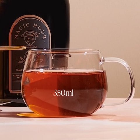 A Kinto Glass Tea Cup filled with 350ml of organic tea sits on a table against a neutral background. Behind the cup, there is a bottle labeled "Magic Hour - Timeless." The scene is well-lit, highlighting the tea's amber color.