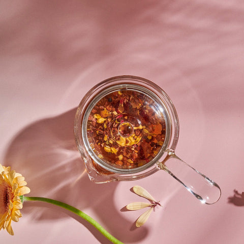 A **Tea-in-Hand: The Perfect Steep Side-pour Ceremonial Teapot** by **Espresso Parts** made of borosilicate glass filled with floral loose leaf teas is placed on a pink surface with a daisy and a decorative model of a dragonfly nearby. The shadows of the daisy and dragonfly are visible, adding a whimsical feel to the scene.