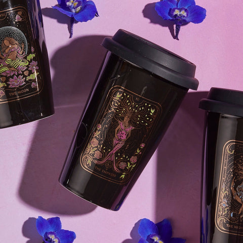 Three black travel mugs featuring tarot card designs—"The Moon," "The Empress," and "The High Priestess"—are arranged on a pink surface. Surrounded by scattered purple flowers, these mugs set the perfect scene for enjoying your The Queen of Cups - Tarot To Go! or Magic Hour in style.