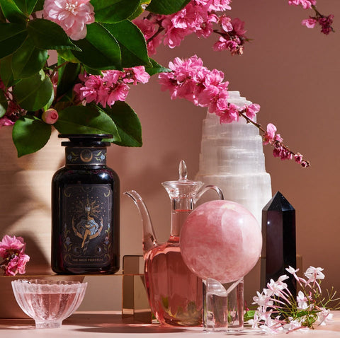 A decorative arrangement featuring a dark glass bottle with ornate design, a clear glass jug of Magic Hour's The High Priestess: Wisdom Tea for Powerful Dreams & Illuminated Insights, a pink crystal sphere on a stand, a white crystal tower, a black crystal obelisk, and a bouquet of pink flowers in a vase, all on a pink surface.