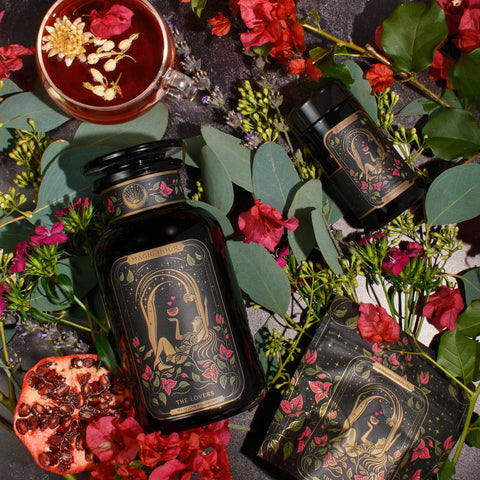 An ornate black jar labeled "The Lovers" from Magic Hour, surrounded by pink and red flowers, eucalyptus leaves, a pomegranate half, a tea cup with floating flowers, a smaller jar, and a box. The setting is a dark surface with a romantic and botanical theme featuring Organic Hibiscus.