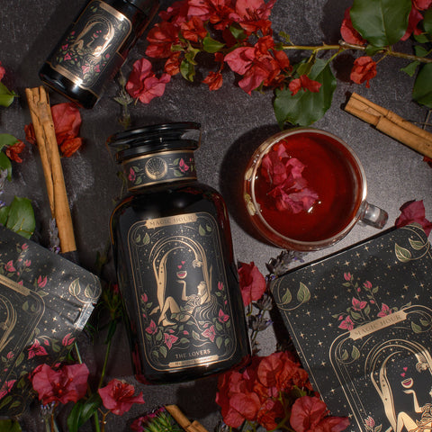 A stylized black bottle labeled "The Lovers" by Magic Hour is surrounded by red flowers, green leaves, cinnamon sticks, and a cup of organic hibiscus tea with flower petals. An accompanying box with mystical illustrations complements the bottle in a sophisticated, nature-themed flatlay.