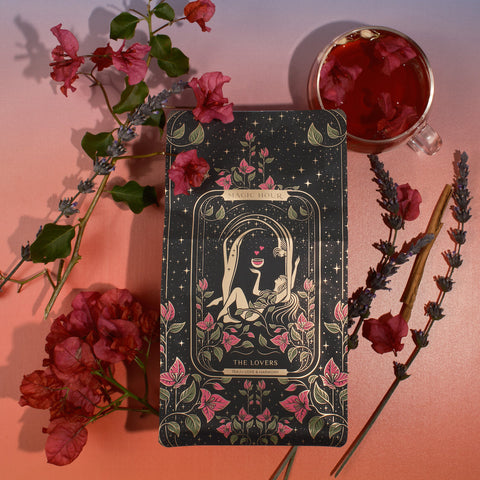 A tarot card titled "The Lovers" is surrounded by decorative flowers, lavender sprigs, and cinnamon sticks on a gradient background. The card features an illustration of two figures intertwined under a crescent moon and stars, with a chalice above them. A cup of Magic Hour's The Lovers tea infused with Organic Hibiscus sits nearby.