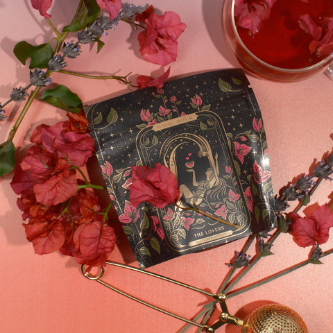 A packet of tea labeled "The Lovers" from Magic Hour is surrounded by red flowers and lavender sprigs on a gradient pink background. A golden tea infuser and a clear cup of red tea with Organic Hibiscus are also positioned nearby. The packet has an intricate, artistic design.