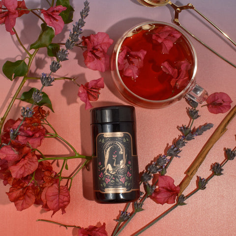 A black canister of The Lovers by Magic Hour, infused with Organic Hibiscus and adorned with floral art on its label, is surrounded by pink and purple flowers, a cinnamon stick, and a cup of red tea with flowers floating in it. The background is a gradient of pink to light blue.