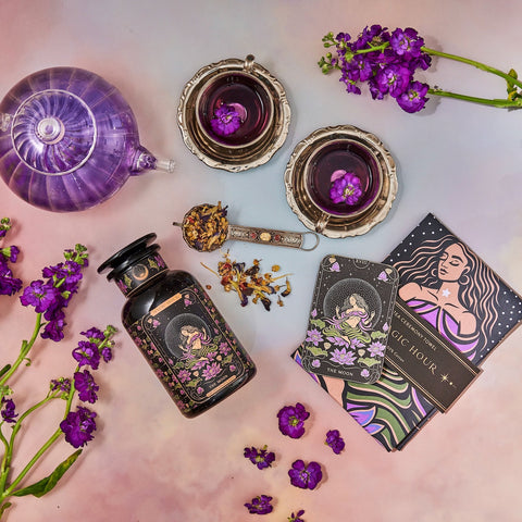 A colorful flat lay features a purple teapot and two teacups filled with herbal tea, surrounded by flowering purple plants. A jar of organic "The Moon - Tea for Nurturing Graceful Phases Case Packs" from Magic Hour, a tarot card labeled "The Moon," and a book titled "Magic Hour" complete the whimsical scene.