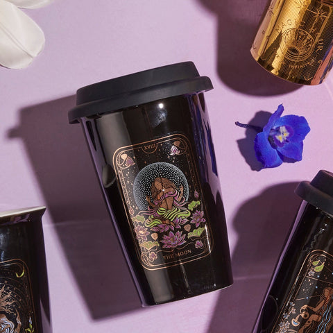 A black reusable cup with a mystical illustration featuring a meditative figure and lotus flowers, labeled "The Queen of Cups - Tarot To Go!," is placed on a light pink surface. The lid is black, and nearby are another cup of Magic Hour Tea, a golden cylindrical object, a white flower petal, and a blue flower.