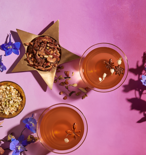 Two glasses of orange-colored "The Star: Vanilla-Ginger Beauty Potion with Jasmine & Shatavari" from Magic Hour, garnished with star anise flowers and other dried botanicals, placed on a pink background. A gold star-shaped container filled with mixed dried botanicals and a small bowl of herbs are nearby. Blue and purple flowers are scattered around.