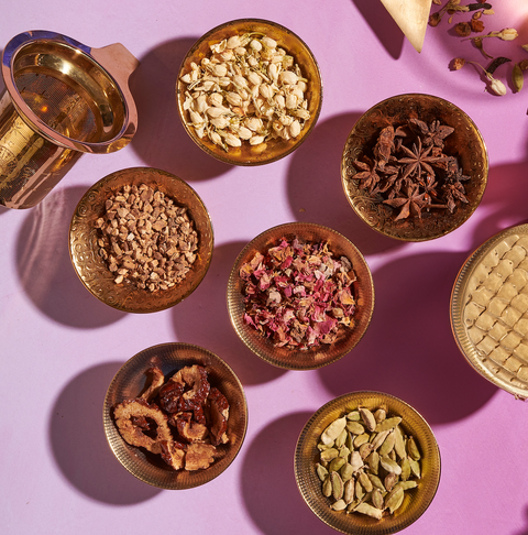 An arrangement of six bowls filled with various dried ingredients on a pink surface. The items include seeds, spices, and dried flowers, perfect for crafting your own loose leaf tea using The Star: Vanilla-Ginger Beauty Potion with Jasmine & Shatavari from Magic Hour. A brass mortar and pestle, along with a triangular cloth bag, are also present in the composition.