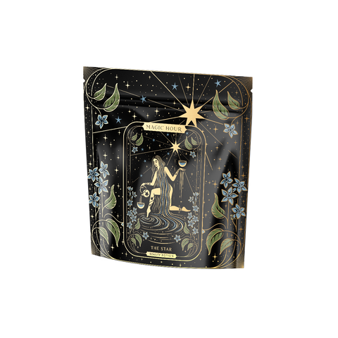 A black tea packaging displaying an intricate illustration of "The Star" tarot card. The design features a woman pouring water, surrounded by a celestial theme with stars and decorative floral elements. Text reads "Magic Hour" and "The Star: Vanilla-Ginger Beauty Potion with Jasmine & Shatavari". Indulge in this organic loose leaf tea for a truly magical experience.