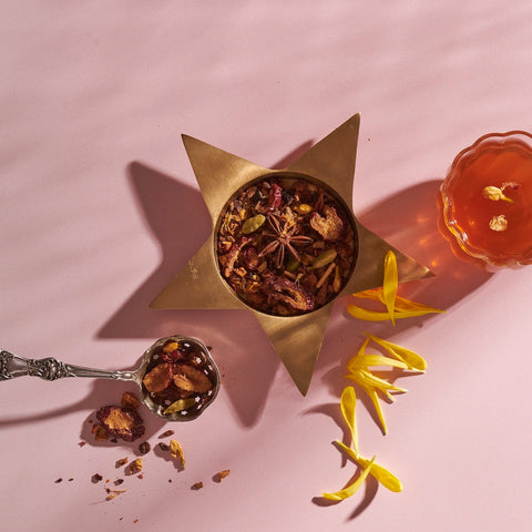 A bowl of Magic Hour’s The Star: Vanilla-Ginger Beauty Potion with Jasmine & Shatavari rests in a star-shaped holder on a pink surface, accompanied by a small glass cup of the tea blend. An ornate spoon holds some loose tea blend with natural flavors. Yellow flower petals are scattered nearby, creating an elegant arrangement.