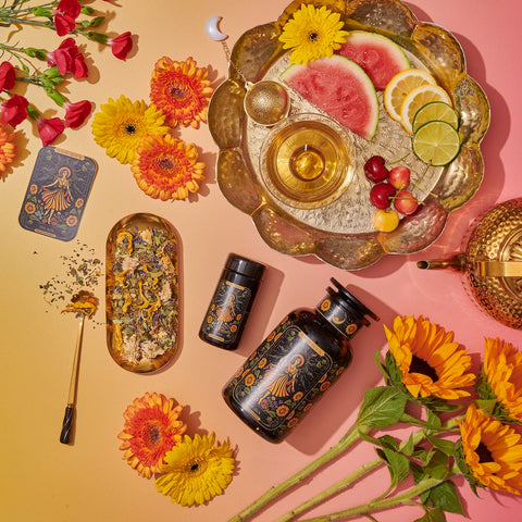 A vibrant flat lay showcases Magic Hour's tea products, "The Sun: Energizing Adaptogenic Herbal Tea with Yaupon, Moringa & Turmeric," in elegant black and gold packaging, surrounded by colorful flowers. A gold tray holds sliced fruits, a tea strainer, and a teacup. Loose Yaupon tea leaves and a golden teaspoon are artfully arranged nearby against a gradient yellow-pink background.