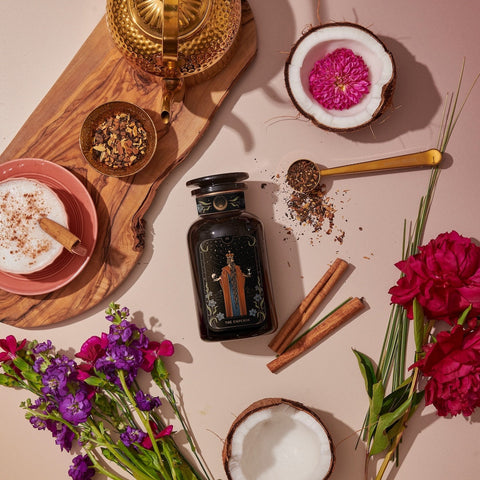 A flat lay image showcasing a bottle of "The Emperor: Green Chaga Chai with Lion's Mane and Reishi," featuring its sleek black design surrounded by fresh flowers, a split coconut, cinnamon sticks, a cup filled with frothy milk, and a wooden tray holding a brass pot along with Gunpowder green tea leaves. A golden teaspoon and scattered tea leaves elegantly complete the scene.