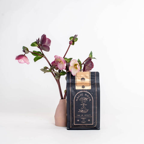 A bag of Magic Hour Ti Quan Yin Oolong: Tea of Patient Compassion stands against a white background. The bag is black with gold details. Beside it, a minimalist vase holds a stem with several purple and pink flowers. This arrangement highlights the tea's wellness benefits and is aesthetically pleasing.