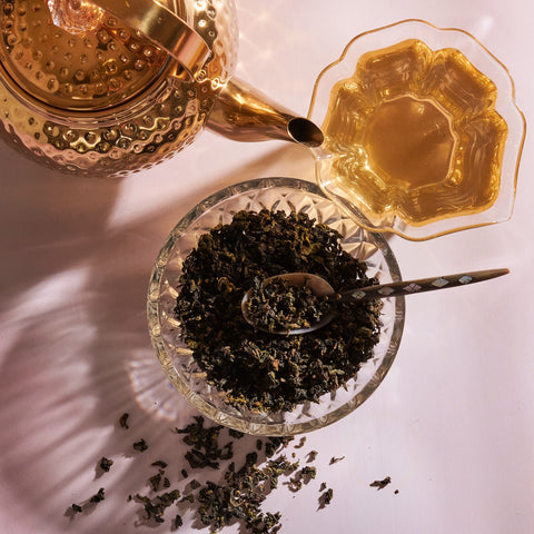 A gold teapot is positioned beside a full, ornate teacup filled with Ti Quan Yin Oolong: Tea of Patient Compassion. In the center, a glass bowl contains loose Ti Quan Yin Oolong: Tea of Patient Compassion leaves with a spoon resting inside. Some loose leaves are scattered around the bowl on the white surface, hinting at their wellness benefits.