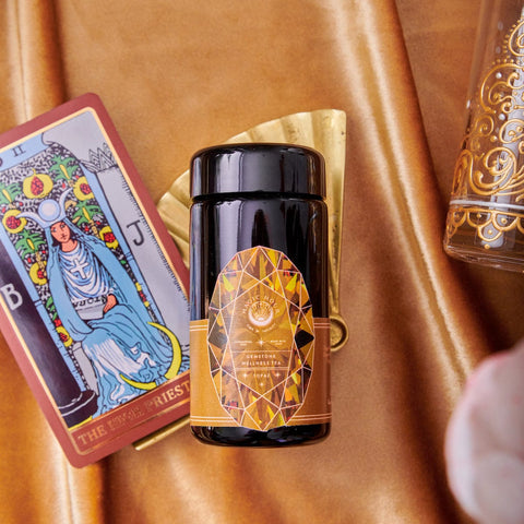 A black jar of Magic Hour’s “Topaz - Caramel Apple Oolong Tea Case Packs” with an intricate, geometric design label sits on a golden surface, perfect for cozy sips. It is accompanied by a tarot card featuring The High Priestess, a golden fan, and part of an ornate glass. The background features a rich, golden-brown fabric texture reminiscent of caramel apple crisp.