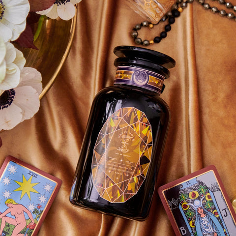 The Topaz - Caramel Apple Oolong Tea Case Packs by Magic Hour, featuring an elegant black bottle with a geometric label, rests on a luxurious silky gold fabric, evoking the feel of cozy sips. Surrounding the bottle are tarot cards, including "The Star" and "The High Priestess." Delicate white flowers and a beaded bracelet further enhance the enchanting scene.