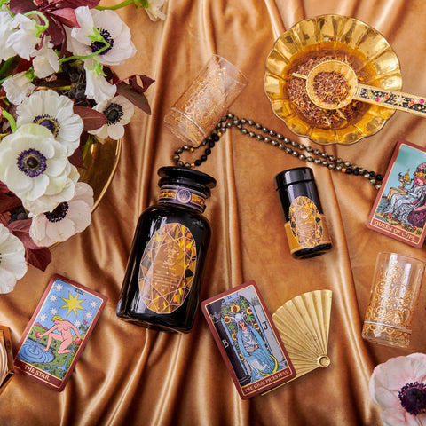 An elegant display featuring tarot cards, a black and gold bottle, glasses, a fan, and a floral arrangement on a silky, golden fabric. The tarot cards are arranged around the bottle, lending a mystical and luxurious ambiance to the setting—perfect for cozy sips of Magic Hour's Topaz - Caramel Apple Oolong Tea from the case packs.