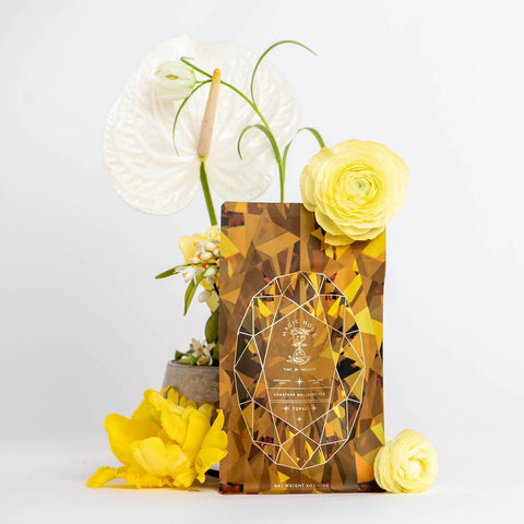 Placed in front of a white background is an elegant case pack of Magic Hour's Topaz - Caramel Apple Oolong Tea. Surrounding the case pack are vibrant yellow flowers and pristine white anthuriums, adding a touch of sophistication and color to the scene, perfect for cozy sips that warm your soul.