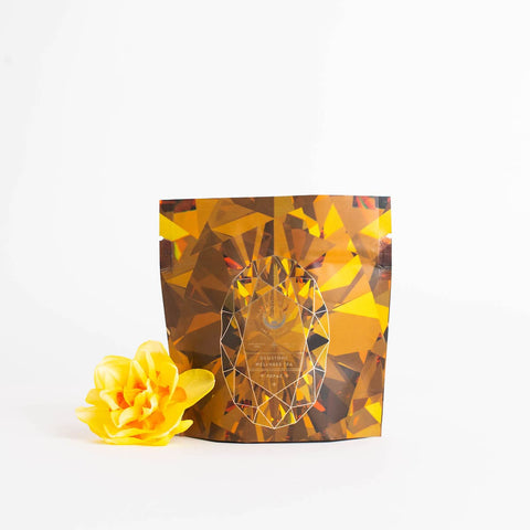 The Topaz - Caramel Apple Oolong Tea Case Packs from Magic Hour feature a multicolored geometric-patterned packaging with gold and brown hues, standing upright against a white background. To the left of the bag is a yellow flower, hinting at the cozy sips within, perfect for enjoying a caramel apple crisp treat.