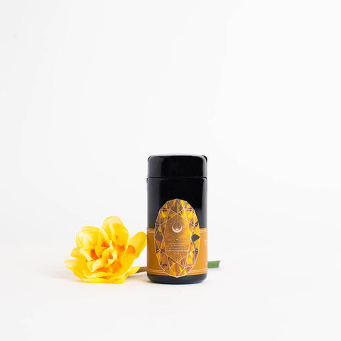 A sleek, black container of Magic Hour's Topaz - Caramel Apple Oolong Tea Case Packs with a golden, geometric design on the label stands against a white background. A vibrant yellow flower rests beside the container, adding a touch of elegance and contrast to the minimalist scene, evoking cozy sips in every detail.