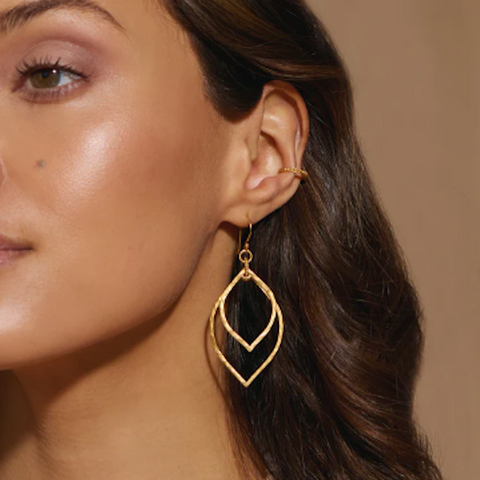 Close-up of a person wearing Magic Hour's Transformed by Light Lotus Linear Earrings with a modern, layered leaf-like design, reminiscent of lotus petals. Their hair is dark brown and wavy, complemented by a small 18kt gold plated ear cuff. The background is a neutral beige color.