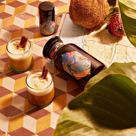 Two cocktails garnished with cinnamon sticks are placed on a patterned tile surface. Next to them is a dark glass bottle of Magic Hour's Tulum Horchata Chai Case Packs, featuring a colorful label and accompanied by a small container, hinting at an adaptogenic wellness potion. The setting includes a coconut, flower, and map partially covered by large green leaves.