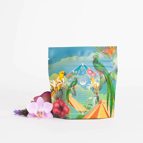 A vibrant packet of Tulum Horchata Chai Case Packs by Magic Hour, adorned with colorful illustrations of birds, flowers, and tropical foliage. The packet stands against a white background with fresh blooms artistically arranged at its base.