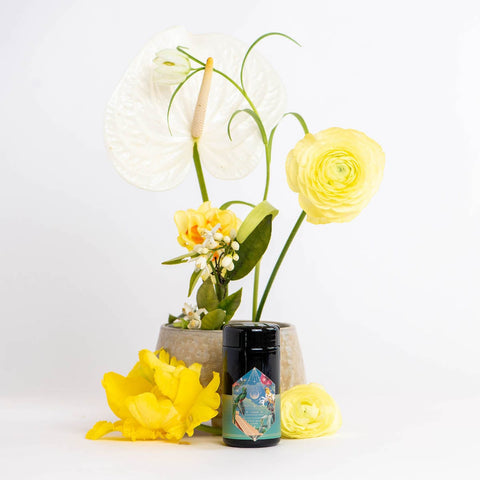 A small jar with a colorful label, featuring Magic Hour's Tulum Horchata Chai Case Packs adaptogenic wellness potion, sits in front of a ceramic pot containing yellow and white flowers, including anthuriums and ranunculus, against a plain white background. Loose yellow and white flowers are scattered around the jar, reminiscent of Tulum's vibrant flora.