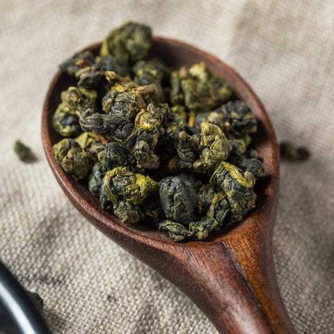 A wooden spoon holds a heap of semi-oxidized oolong tea leaves, displaying a mix of curled and somewhat wavy textures. The spoon rests on a fabric surface, highlighting the rich, dark green and brown tones characteristic of Magic Hour's Tung Ting Green Dragon Oolong from Taiwan.