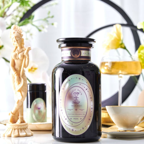 A decorative setup featuring a large dark-colored jar labeled "Pearl Tea of Beauty, Clarity, and a Sense of Peace" from Magic Hour surrounded by luxurious items, including a small bottle, a cup and saucer ready for loose leaf tea, and a delicate statue. Soft lighting and flowers create an elegant, serene atmosphere.