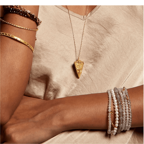 A person wearing a beige top is shown in partial view. They have multiple gold and beaded bracelets on their left wrist, 18kt gold bangles on their right arm, and the Divine Messages Pendulum Locket Necklace featuring a triangular gold pendant. Their arms are crossed in front of them.