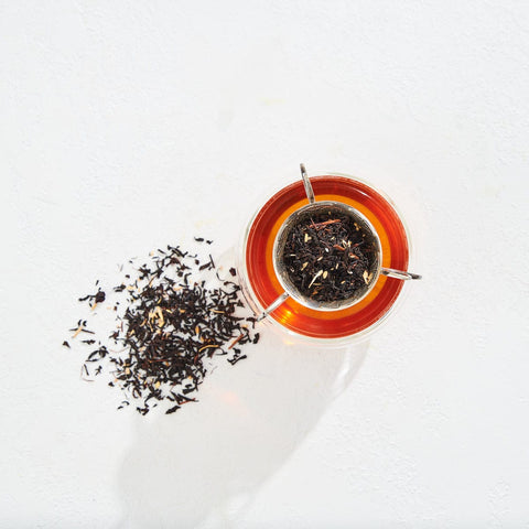 A cup filled with amber-colored Virgo Tea of Virtue, Wit & Meticulous Magic sits on a white surface with a tea strainer containing loose tea leaves resting on top. To the left of the cup, a small pile of loose leaf tea is scattered on the surface. The scene is brightly lit, evoking a serene Magic Hour moment.