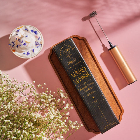 A Wand Whisk: Rechargeable Stainless Steel Eco-Frother, a whisk, a wooden box labeled "Wand Whisk," and a ceramic teacup with white frothy liquid topped with blue flower petals are arranged on a pink surface. Surrounded by white baby's breath flowers, this delicate scene invites you to enjoy the enchantment of Magic Hour Tea.