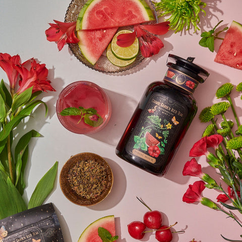 A jar of Watermelon Mint Herbal Iced Tea is surrounded by a glass of iced tea, sliced watermelon, cherries, lime slices, red flowers, and a bowl of loose tea leaves with a hint of organic peppermint. The setup is laid out on a white surface.