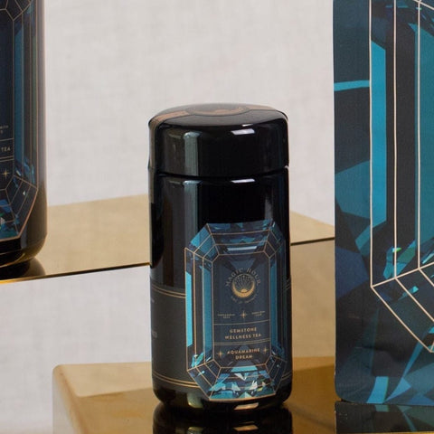 A black cylindrical container with a blue geometric design sits on a golden reflective shelf. The label reads "Magic Hour" with "Aquamarine Dream - Soothing Herbal Ayurvedic Adrenal Tonic" written below. This loose leaf tea is flanked by a similar design on the right, adding to the allure of Magic Hour Tea.