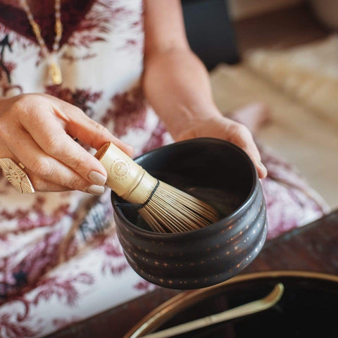 A person is using a wooden whisk to froth Magic Hour Tea in an Alibaba Ceremonial Tea Bowl. The individual is wearing a patterned garment and a gold ring, with part of a gold necklace visible. A tray with other utensils is in the background.