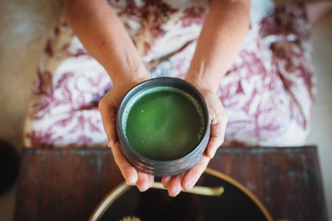 A person holding a black ceramic Ceremonial Tea Bowl filled with vibrant green Magic Hour Tea matcha. The person is seated and wearing a patterned garment with hues of red and beige. The background features a wooden surface and partial view of another black bowl from Alibaba.