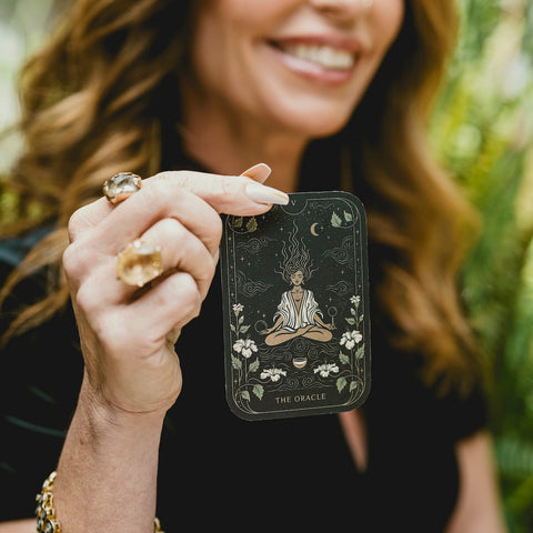 A woman with wavy hair holds up a tarot card titled "The Oracle." The card features an illustration of a figure with flowing hair, sitting in a meditative pose, surrounded by lilies, with celestial symbols above. The woman smiles, with greenery in the blurred background.

Magic Hour