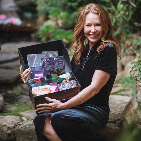 A woman with long, wavy, auburn hair, wearing a black dress, smiles while holding an open black box filled with the latest delights from her Tea School Course #1: Tea Essentials + Taste New Teas by Magic Hour. She is seated on a stone surface in an outdoor, garden-like setting with greenery in the background.