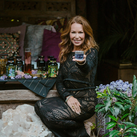 A woman with long, wavy hair, dressed in a black lace outfit, sits on a wooden bench. She smiles and extends a glass of floral tea towards the viewer. The background has a cozy setup with decorative pillows, jars, flowers, and greenery, creating a luxurious and inviting atmosphere—perfect for showcasing Magic Hour's Tea School Course #5: Tisanes - Herbs, Medicinals, Mocktails & Flowers.