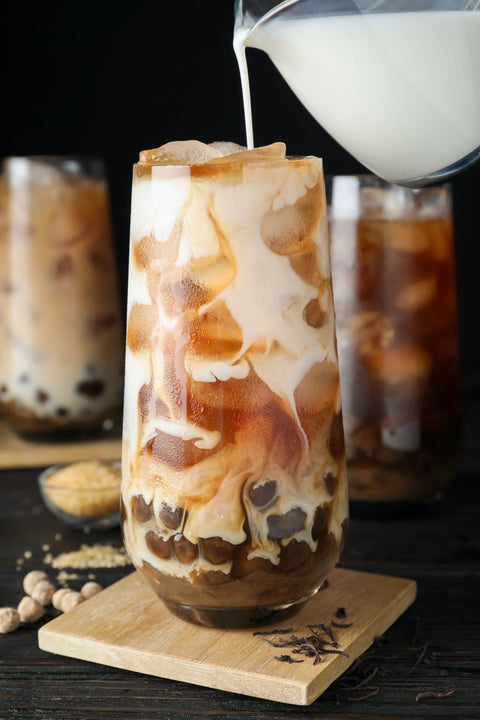 A glass filled with iced bubble tea made with Boba: Tapioca Pearls by Magic Hour, with milk being poured into it. The swirling effect of the milk mixing with the organic tea creates a marbled appearance. Two other glasses of bubble tea made with Boba: Tapioca Pearls by Magic Hour are blurred in the background, along with a small bowl containing ingredients.