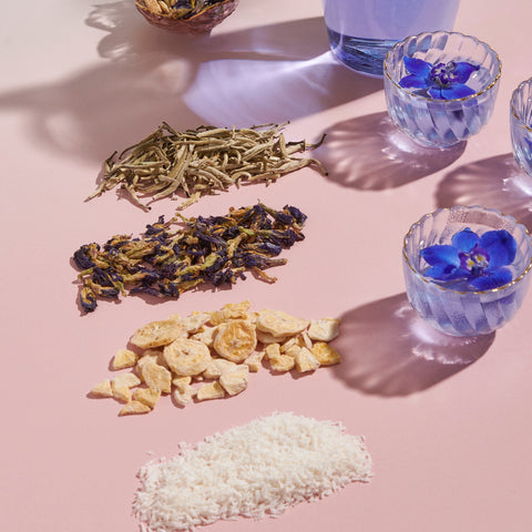 An arrangement of dried tea leaves, dehydrated fruit pieces, and shredded coconut displayed on a pink surface, with three glass cups of Magic Hour Butterfly Blue Coconut Cream Iced White Tea, one containing fresh butterfly pea flowers. Shadows cast over the scene from lighting create a refreshing summer hydration tableau.