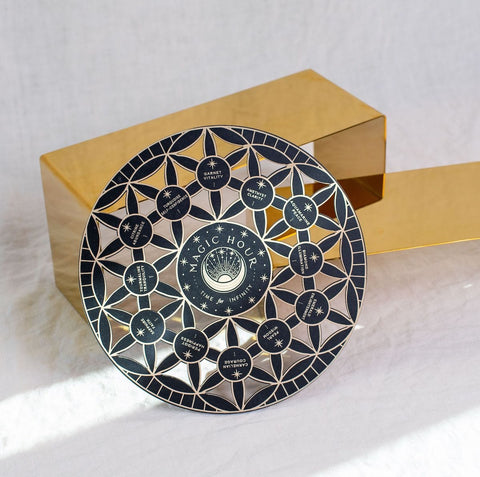 A round black and silver decorative plate with intricate geometric patterns and text "Crystal Grid: Flower of Life Tea Ceremony Altar" at the center, surrounded by smaller circles with text like "gratitude," "immunity," and "healing." The plate is set against a white cloth backdrop, next to a golden rectangular object.